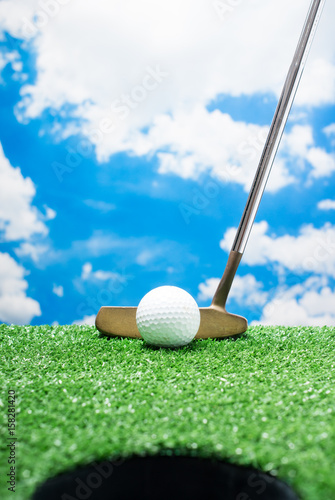 Look through the hole to the golf ball and the putter on fake green grass against cloud and blue sky