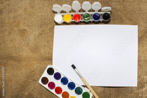 Set of watercolor paints, brushes for painting and blank white paper sheet of sketchbook.
