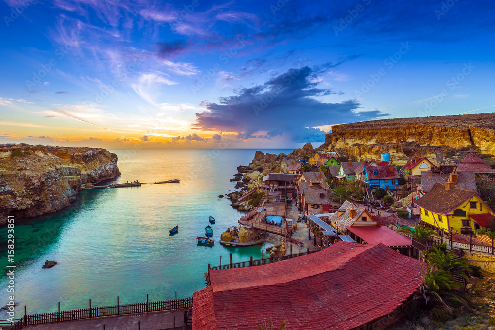 Mellieha, Malta - Skyline view of the beautiful Popeye Village at Anchor Bay at sunset with amazing colorful clouds and sky - obrazy, fototapety, plakaty 