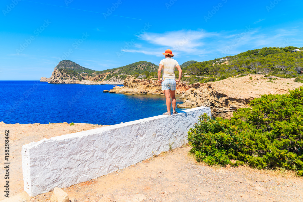 Young woman tourist standing on white wall and looking at  Punta Galera bay surrounded by amazing stone formations, Ibiza island, Spain