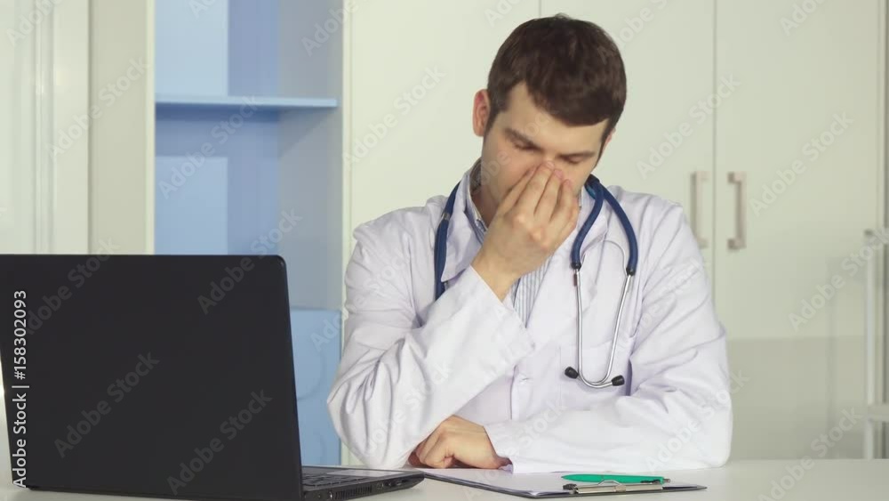 Handsome male doctor getting tired at the office. Attractive caucasian man in white coat massaging his temples. Brunette medical specialist finishing to work on laptop Handsome male doctor getting tired at the office. Attractive caucasian man in white coat massaging his temples. Brunette medical specialist finishing to work on laptop