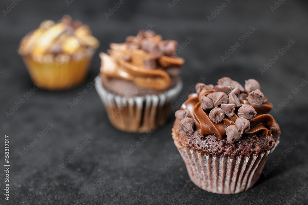 Three chocolate cupcakes garnished with chocolate chips and chocolated frosting on lined up across the frame, on black background, from left to right Three chocolate cupcakes garnished with chocolate chips and chocolated frosting on lined up across the frame, on black background, from left to right