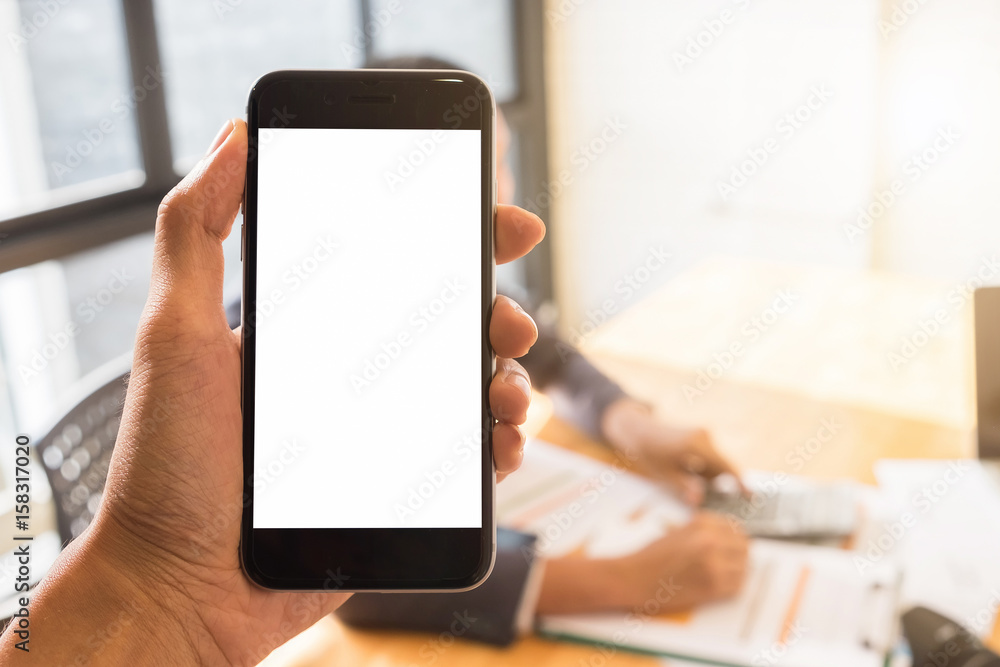 Close up of a man holding smart phone with blank screen mobile and laptop computer with business working in office background.