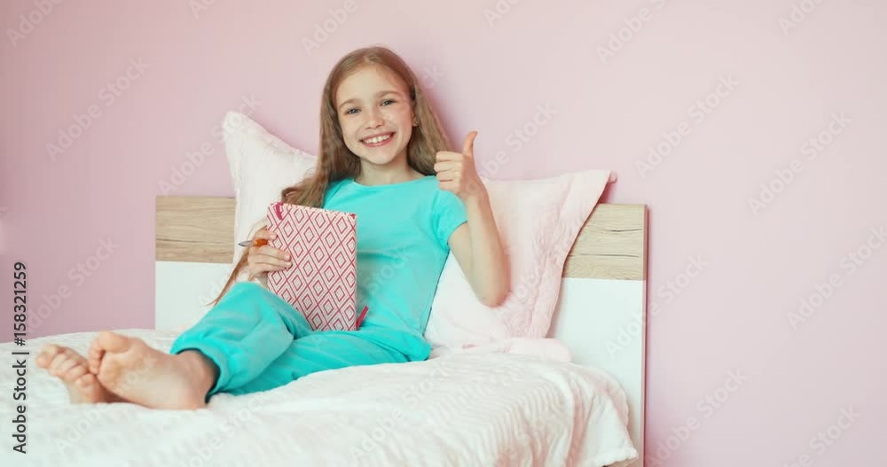Child student sitting on the bed and laughing at camera. Girl holds book Child student sitting on the bed and laughing at camera. Girl holds book