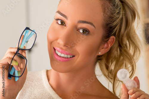 Smiling young blonde woman holding contact lens case on hand and holding in her other hand a blue glasses on blurred background., eyesight and eyecare concept