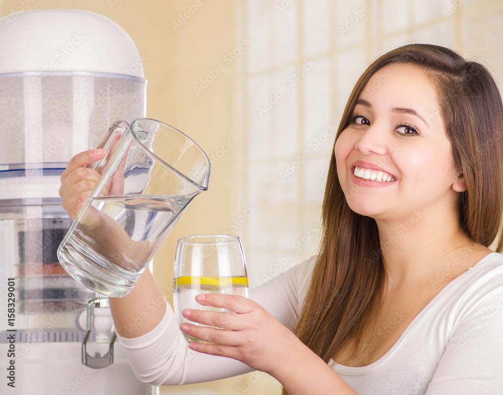 Beautiful smiling woman holding a glass of water in one hand and a pitcher of water in her other hand, with a filter system of water purifier on a kitchen background