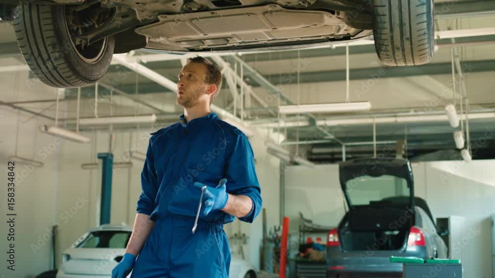 Caucasian young auto mechanic man in blue uniform whistling and smiling. Auto mechanic using wrench standing at autoservice. Indoor. Caucasian young auto mechanic man in blue uniform whistling and smiling. Auto mechanic using wrench standing at autoservice. Indoor.