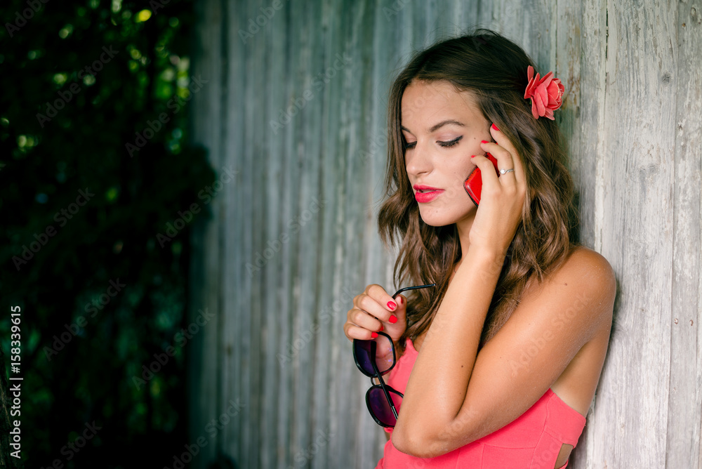 Obraz premium Young sexy stylish lady talking on mobile phone in front of wooden door on sunny summer day. Lifestyle portrait outdoor background