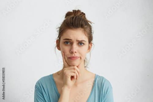 Photography Young woman with fair hair and blue eyes frowning her eyebrows holding index finger on chin having doubt and suspicion feeling sceptical about something