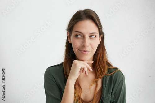 Headshot of cunning beautiful woman with straight dyed hair looking aside holding hand under chin indenting to realize tricky plan. Attractive female having sly expression while looking aside