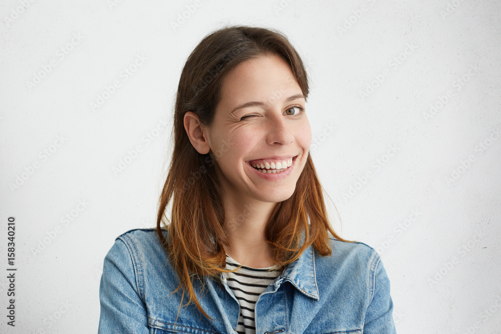 Indoor portrait of beautiful girl dressed in denim jacket over striped top smiling broadly and winking mysteriously at camera, having playful look. Positive human facial expressions and emotions Indoor portrait of beautiful girl dressed in denim jacket over striped top smiling broadly and winking mysteriously at camera, having playful look. Positive human facial expressions and emotions