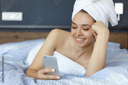 Pleased woman with white towel on head lying in bed relaxing after taking shower holding smartphone messaging in social networks before sleep having happy expression on face. Relief after hard work