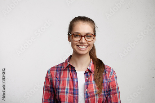 Cute female with green eyes and long eyelashes in big round eyeglasses and red checked shirt looking elegant having pleasant broad smile isolated over white background. Beauty and youth concept © wayhome.studio 