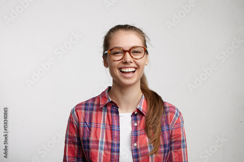 Adorable female in round spectacles and red checked shirt having fair hair tied in pony tail looking with excitement into camera having white even teeth. People, sincere emotions, lifestyle concept © wayhome.studio 