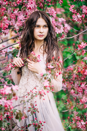 beautiful sexy girl in delicate lace dress beige on a background of pink Sakura, hands touching twigs looks into the distance photo