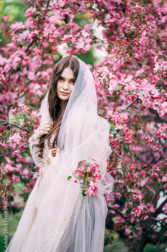 beautiful sexual girl brunette bride in long wedding dress beige, closed the veil to the hair and looks with a smile, happy, on the background of pink cherry blossoms, tree, nature, Park