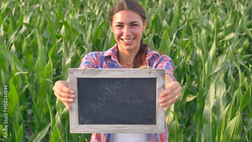 Beautiful girl (woman) farmer smiling, looking, checking cornfield, holding a black board, background of greenery. Concept: ecology, corn, bio product, inspection, water, natural products, environment photo