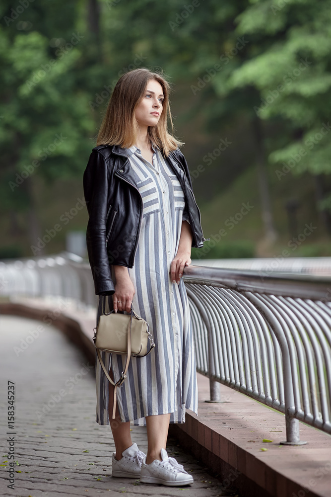 Young beautiful girl in stylish streetwear black leather jacket long striped dress white sneakers and with a fashionable bag strolling in summer city park