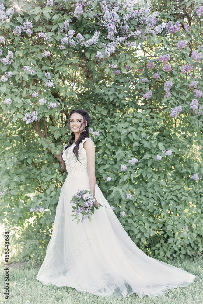 Tender beautiful cute girl bride in a white air dress with a bouquet of lilacs in her hands walking through the park on a sunny spring day. Photo in gentle colors
