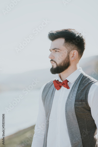 Portrait of stylish groom man in white shirt, gray waistcoat and red bow-tie at the wedding in nature forest and seacoast. Selective focus. Toned image