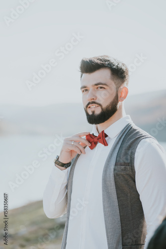 Portrait of stylish groom man in white shirt, gray waistcoat and red bow-tie at the wedding in nature forest and seacoast. Selective focus. Toned image