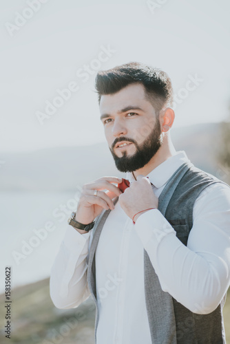 Portrait of stylish groom man in white shirt, gray waistcoat and red bow-tie at the wedding in nature forest and seacoast. Selective focus. Toned image