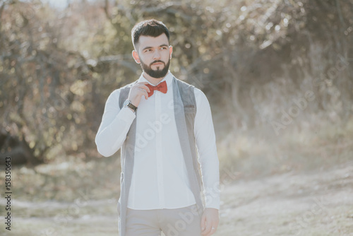 Portrait of stylish groom man in white shirt, gray waistcoat and red bow-tie at the wedding in nature forest. Selective focus. Toned image