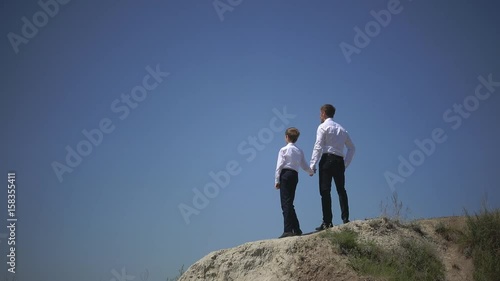 Father and son are standing on the hill holding hands and looking confidently into the distance, dreaming about the happy future of the family, against the background of the blue sky,