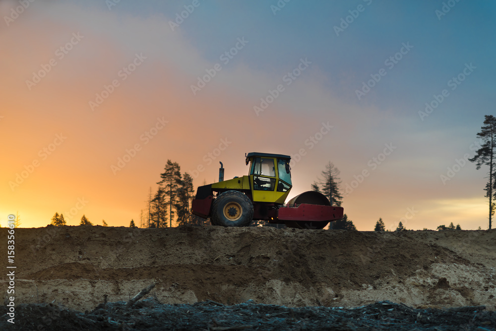 Road roller at road construction site with yellow and blue sky during sunset Road roller at road construction site with yellow and blue sky during sunset