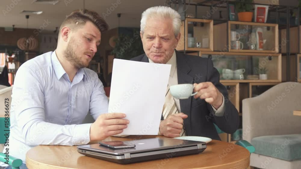Two male business partners discussing some papers at the cafe. Young bearded man turning pages of some document. Senior caucasian businessman drinking tea near his colleague Two male business partners discussing some papers at the cafe. Young bearded man turning pages of some document. Senior caucasian businessman drinking tea near his colleague
