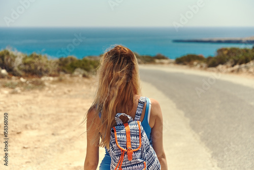 Beautiful young girl walking up to the sea along the road. She has rucksack on her back.
