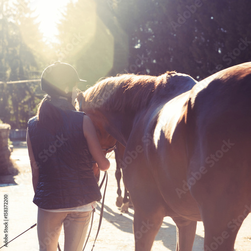Young teenage girl equestrian leading her brown horse in sunbeam