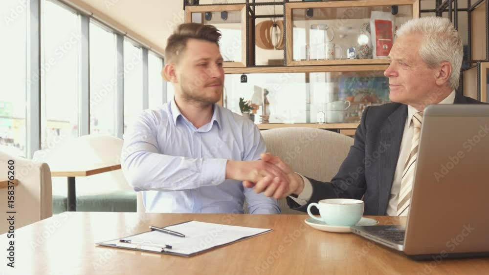 Two caucasian business partners shaking hands at the cafe. Young bearded guy sitting down near his older companion. Attractive blond man pointing his pen on clipboard with papers