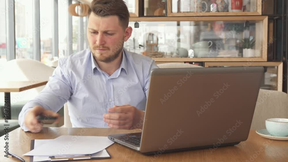 Young bearded businessman calling on the phone at the cafe. Attractive blond man turning pages of some document. Handsome caucasian guy dressed in white shirt taking smartphone from the table Young bearded businessman calling on the phone at the cafe. Attractive blond man turning pages of some document. Handsome caucasian guy dressed in white shirt taking smartphone from the table