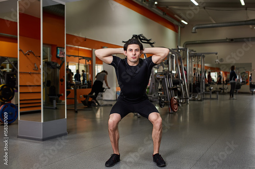 Portrait of young sportsman in the black sportwear while making squat in gym.