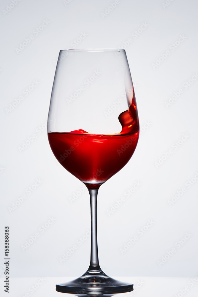 Splashing and moving red wine in the pure wineglass standing against light background with reflection on the stand.