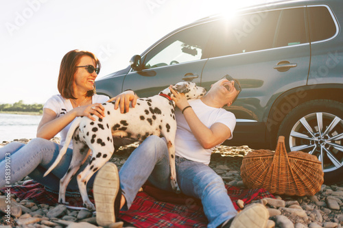 Portrait of happy young adult couple with dog on roadtrip. Man sitting on plaid with woman. Outdoor picnic concept.