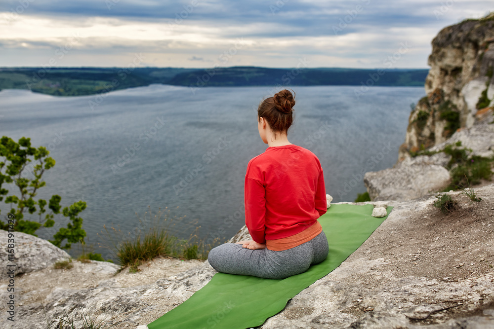 Rear view of young woman sitting in lotus position on the rock over the river.