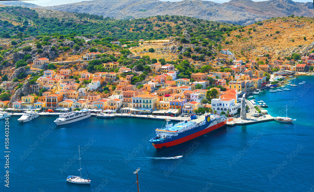 View on beautiful traditional Greek houses on Symi island green hills, yacht sea port, tourist ferryboat at Aegean Sea blue bay. Mediterranean MSC cruises. Greece islands holiday vacation tours trip
