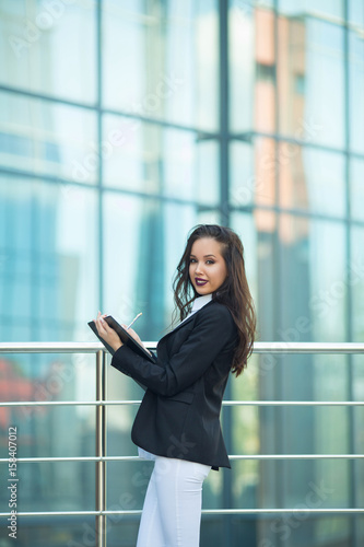 Beautiful young girl in a black jacket with a folder and pen in hand on the background of a glass building
