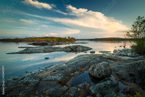 Summer sunset with reflection in water. The clouds are reflected in the water. Stones on the beach. White Nights. Karelia. Ladoga lake.
