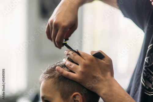 Little Boy Getting Haircut By Barber While Sitting In Chair At Barbershop. 