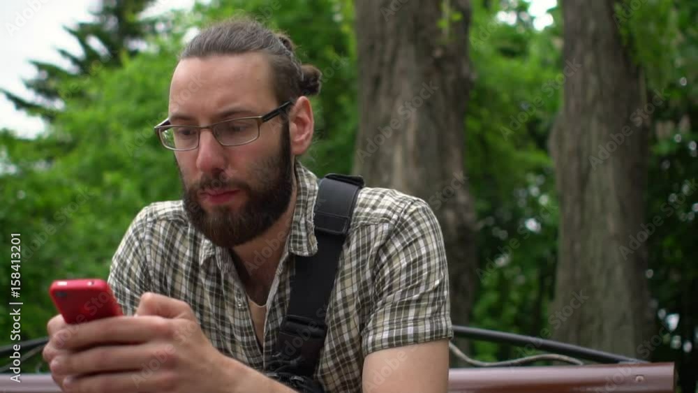 Handsome man with beard and glasses typing text in a park on his smartphone Handsome man with beard and glasses typing text in a park on his smartphone
