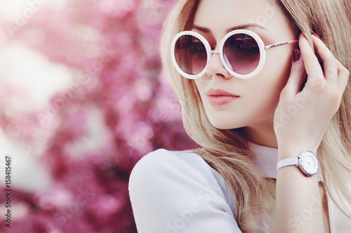 Outdoor close up portrait of young beautiful fashionable girl posing in street. Model wearing stylish white round sunglasses, wrist watch. Female fashion concept. Copy, empty space for text. Toned