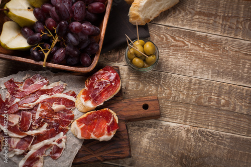 Fototapeta Naklejka Na Ścianę i Meble -  Jamon Iberico with white bread, olives on toothpicks and fruit on a wooden background. Top view with copy space