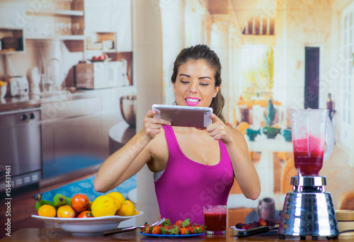 Young woman wearing pink top enjoying healthy breakfast, eating fruits, taking picture using mobile phone and smiling, home kitchen background