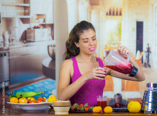 Young woman wearing pink top enjoying healthy breakfast, eating fruits, using blender preparing smoothie and smiling, home kitchen background