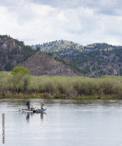 Two fishermen and a third man rowing a boat in the Missouri River in Montana. Willows along the river bank and foothills are seen in the distance and cloudy sky above.