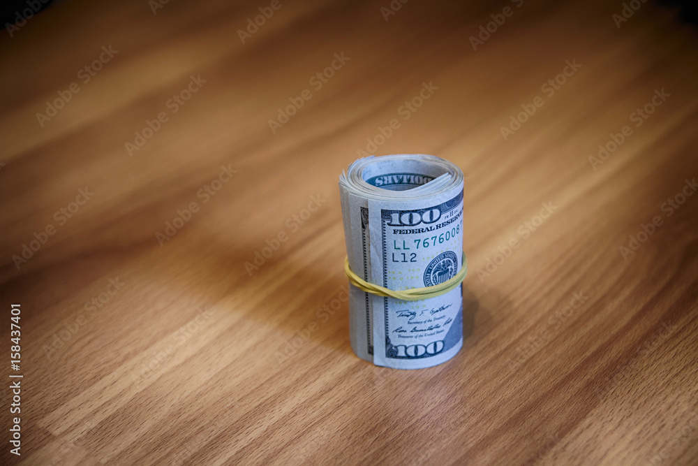 A roll of hundred-dollar bills stitched with rubber band stands on a wooden table A roll of hundred-dollar bills stitched with rubber band stands on a wooden table
