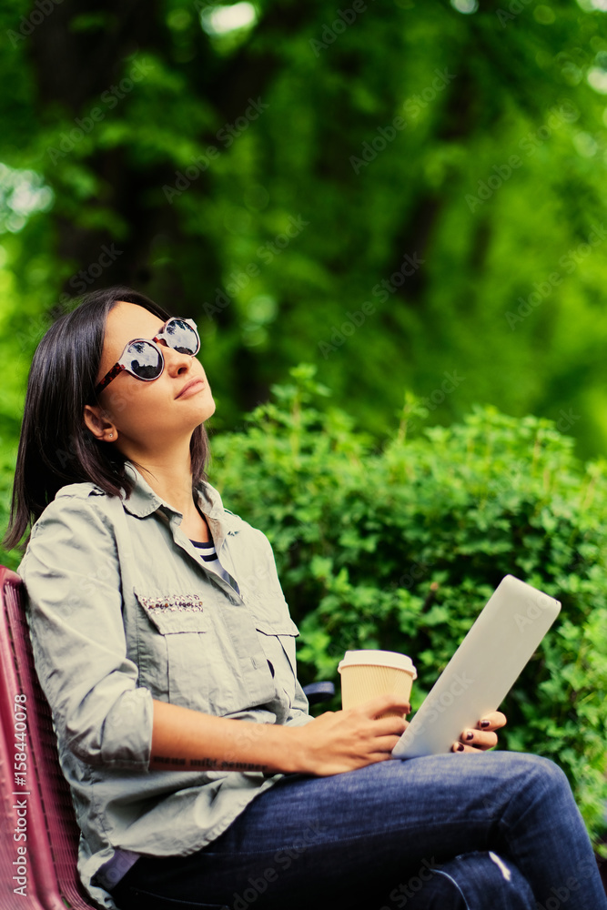 Brunette female holds tablet PC drinks coffee in a summer park Brunette female holds tablet PC drinks coffee in a summer park